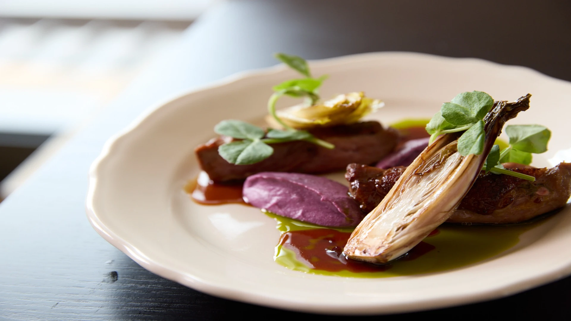 Plated gourmet dish featuring glazed meat, a roasted endive, and a dollop of purple purée, garnished with microgreens and sauce on a cream-colored plate