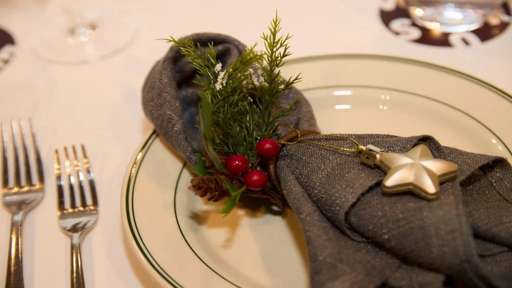 Place setting with a gray napkin wrapped in a ring, decorated with evergreen sprigs, red berries, and a small gold star ornament on a plate beside two forks.