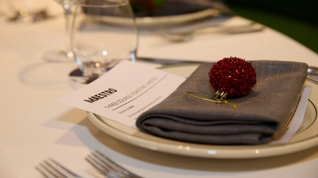 Place setting with a printed “MAESTRO” three-course feasting menu on a plate, two forks beside it, and a loaf of bread on a white tablecloth.