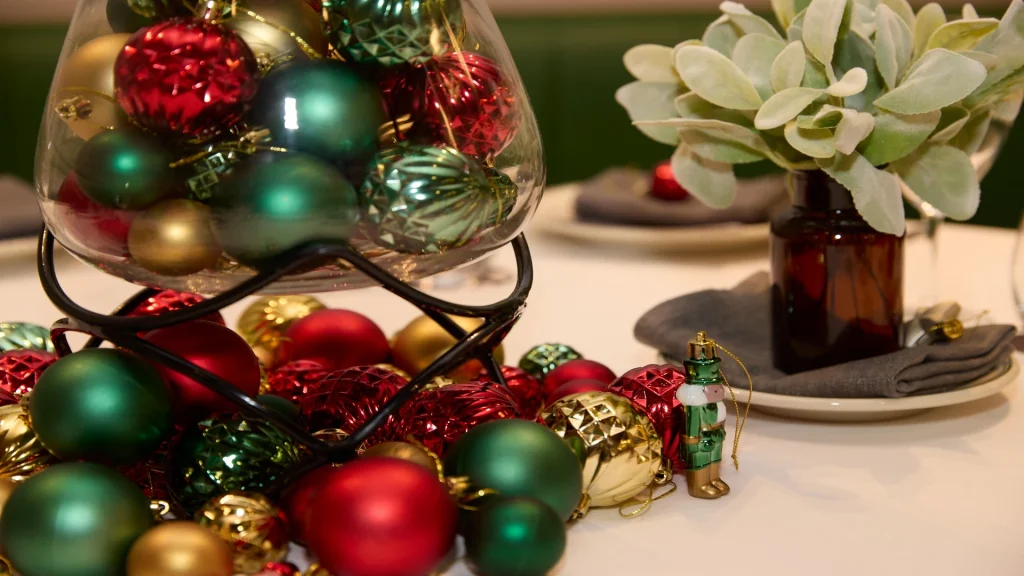 Close-up of a holiday table centerpiece with red, green, and gold ornaments in a glass bowl, surrounded by more baubles, a small nutcracker figurine, and a vase of greenery.
